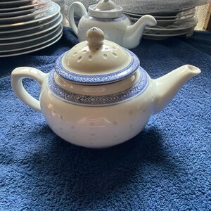 May include: A white ceramic teapot featuring a blue geometric pattern around the rim and lid. The lid has a perforated design with a small knob handle. The body of the teapot is round with a curved spout and a handle on the opposite side.