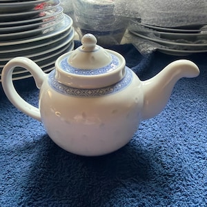 Op de afbeelding: Een witte porseleinen theepot met een blauw geometrisch patroon rond het deksel en de bovenrand. Het lichaam van de theepot heeft een subtiele textuur met reliëfstippen. De theepot is voorzien van een gebogen handvat, een tuit en een bijpassend deksel.