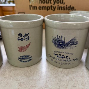 Pode incluir: Dois potes de grés cinza-claro. O pote da esquerda apresenta o número 25 em azul, um logotipo de asa vermelha e um carimbo oval azul com a inscrição Red Wing Stoneware Co. Red Wing, Minn. O pote da direita mostra uma ilustração azul.