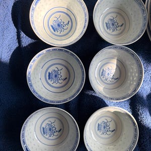 May include: A set of six white porcelain bowls featuring a blue floral design at the centre and a blue geometric pattern along the inner rim. The exterior of each bowl displays a translucent rice grain pattern, creating a delicate texture.