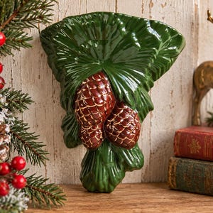 May include: A glossy ceramic wall pocket shaped like a cluster of green pine needles with two brown pinecones in the center. The piece features a deep green glaze on the foliage and a textured brown finish on the pinecones, designed for wall mounting.