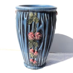 May include: A blue ceramic vase with vertical ribbed detailing. The vase features a floral design of red roses with green leaves. The vase has a slightly flared top and a rounded base. The overall design is elegant and decorative.