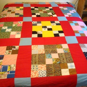 May include: A handmade quilt with a patchwork design. The quilt features a red border and squares of various fabrics in colors like blue, green, yellow, and pink. The quilt is spread across a bed.