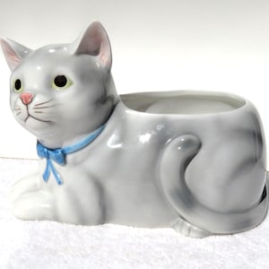 May include: A ceramic cat-shaped planter featuring a glossy light gray finish with darker gray shading. The cat has yellow eyes, a pink nose, and a bright blue painted bow around its neck. The hollow opening for plants is located on the cat's back.