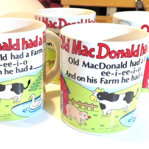 May include: Four white ceramic mugs with the text "Old MacDonald had a Farm" in red and black. Each mug features a farm scene with a cow, pig, duck, and red barn on a green background.