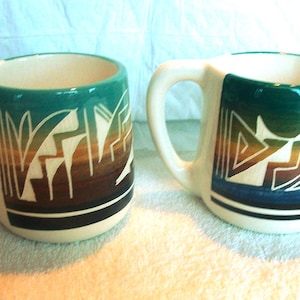 May include: Two ceramic mugs with a cream-colored base and handles. Each mug features a geometric design in shades of teal, brown, and white. A black stripe accents the bottom. The mugs are suitable for coffee, tea, or other beverages.