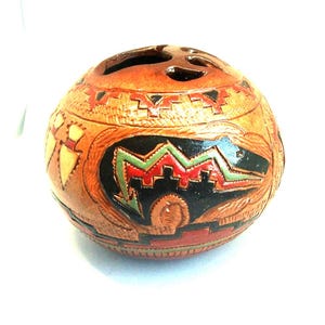 May include: A round, brown, carved decorative object with a series of holes on top. The object features a black, red, and green design on the front. The design appears to be a stylized bear or animal figure. The object is likely made of wood or a similar material.