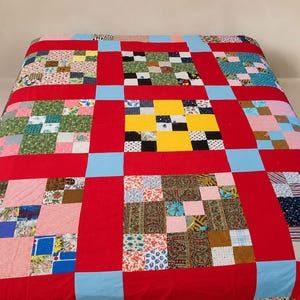 May include: A patchwork quilt featuring a grid of square blocks in various floral, geometric, and solid patterns. The blocks are separated by wide red fabric sashing and accented with light blue squares at the intersections of the grid.