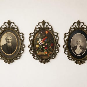 May include: Three oval wall frames with ornate, antique-style bronze-colored metal borders. The left frame contains a sepia-toned portrait of a bearded person, the center frame displays a colorful floral still life, and the right frame holds a black and white portrait of a person wearing a collared garment.