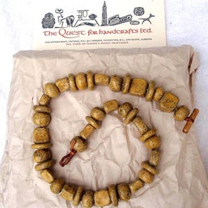 May include: A necklace made of light brown, irregularly shaped beads. The beads are strung together with a brown cord and secured with a wooden toggle clasp. The necklace rests on a piece of brown paper, with a card that reads "The Quest for handcrafts ltd."