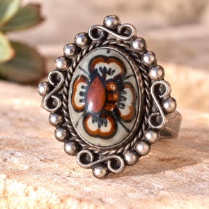 May include: A silver ring featuring an oval ceramic cabochon with a hand-painted floral motif in shades of rust, black, and cream. The setting is decorated with a beaded border and small silver scrolls, creating a detailed frame around the center.