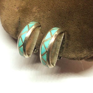 May include: A pair of silver-toned hoop earrings with a turquoise inlay. The turquoise is arranged in a geometric pattern. The earrings are displayed on a brown surface.