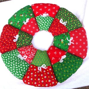 May include: A colorful, handmade quilted wreath with a central white circle. The wreath is composed of wedge-shaped fabric pieces in red and green, each patterned with small white flowers. White yarn bows accent the seams.