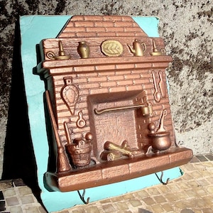 May include: A bronze-colored, decorative fireplace wall hanging with a brick design. The fireplace features a mantel with miniature kitchen tools, a kettle, and a decorative plate. A broom, bucket, and logs are below the fireplace opening. Two hooks are at the bottom.