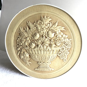 May include: A beige, circular decorative plate with a raised relief design. The central image is a detailed arrangement of flowers in a decorative vase. The flowers include roses, daffodils, and other blossoms. The plate has a smooth, light-colored border.
