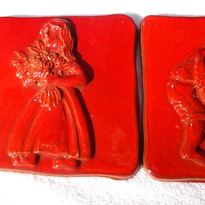May include: Two red ceramic tiles with raised relief designs. One tile depicts a person holding flowers, the other shows two people. The tiles are square and have a glossy finish. The figures are in a folk art style.