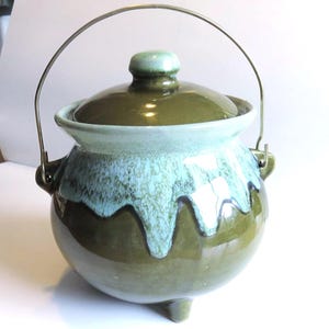 May include: A vintage ceramic cooking pot with a lid and a metal handle. The pot is a rounded shape with three small feet. The color scheme is a mix of olive green, light blue, and brown. The lid has a small knob on top.