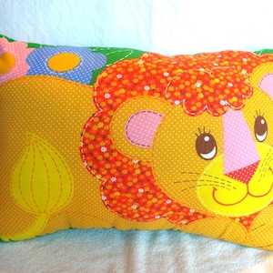 May include: A rectangular pillow featuring a friendly lion design. The pillow has a yellow background with white polka dots. The lion's mane is orange and red, and the face has pink and brown accents. A flower and leaf are also on the pillow.