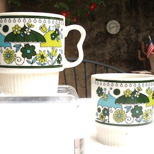 May include: Two white ceramic mugs with a colorful floral and umbrella design. The mugs have a green, blue, and yellow color scheme. The mugs are stacked on top of a white base.