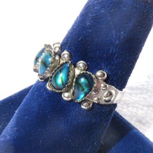 May include: A silver ring featuring three teardrop-shaped iridescent abalone shell inlays with blue and green hues. The metal band is decorated with small silver spheres and a textured finish surrounding the stones in a cluster arrangement.