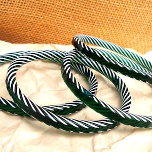 May include: Four green and white striped bangles are displayed on a crumpled paper surface. The bangles are made of a transparent material, with a repeating pattern of diagonal white stripes against a green background. The background includes a burlap texture.
