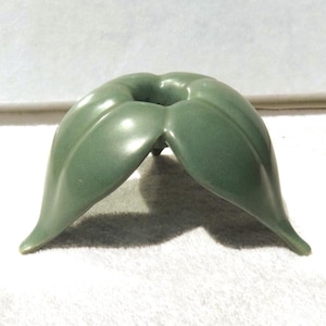 May include: A sage green ceramic candle holder shaped like a stylized flower with four curved petals extending outward from a central circular opening. The object has a smooth, matte finish and rests on the tips of its petal-like base structure.