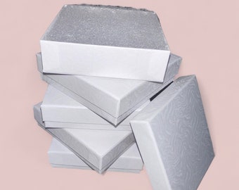 May include: A stack of square silver jewelry boxes with a subtle, swirling embossed pattern on the lids. The boxes are arranged in a vertical pile, with one box standing upright on the side, showcasing the metallic finish and textured surface.