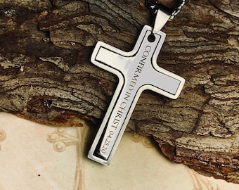 Puede incluir: Colgante de cruz plateada con la inscripción "CONFIRMED IN CHRIST 04.25.20" en una cadena negra. La cruz se encuentra sobre un fondo marrón texturizado.
