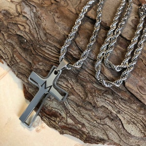 May include: A silver chain necklace with a double cross pendant. The cross is made of two layers of metal, with the top layer cut out to reveal the layer underneath. The cross has a small letter "M" engraved on it.