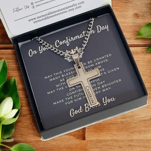 Puede incluir: Un collar con una cruz de plata presentado en una caja negra. La cruz está grabada con el nombre "Cameron". La caja tiene el texto "On Your Confirmation Day" y un mensaje religioso.