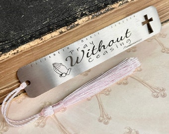 Könnte beinhalten: Silbernes Lesezeichen mit einem rosa Quasten. Das Lesezeichen hat oben ein Kreuz und die Worte "Beten ohne Unterlass 1 Thessalonicher 5,17" sind darauf graviert. Das Lesezeichen hat auch ein Design mit betenden Händen.