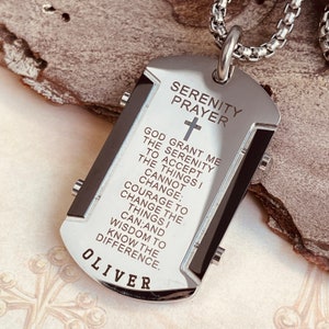 May include: A silver dog tag necklace with a black inlay. The tag features the Serenity Prayer engraved in white text, along with a cross and the name "Oliver".