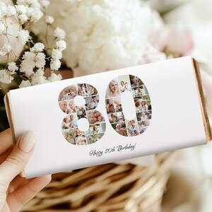 May include: A white rectangular gift box featuring the number "80" made up of various photos. The text "Happy 80th Birthday!" is printed below. The box is held in a hand, with flowers and foliage in the background.