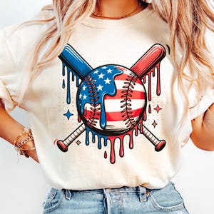 K&ouml;nnte beinhalten: Cremefarbenes Sweatshirt mit einem Baseball-Motiv im rot-wei&szlig;-blauen Design der amerikanischen Flagge. Zwei gekreuzte Baseballschl&auml;ger, einer blau und einer rot, befinden sich hinter dem Baseball. Das Design hat einen tropfenden Farbeffekt.