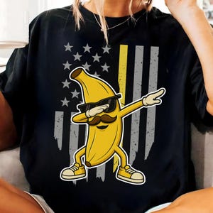 May include: Black t-shirt featuring a yellow banana character with sunglasses and a mustache, dabbing. The design incorporates an American flag with stars and stripes, including a yellow stripe. The banana is wearing yellow sneakers.