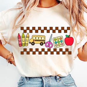 May include: Cream-colored sweatshirt featuring a school-themed graphic. The design includes smiling crayons, a yellow school bus, pink scissors, a tray of smiley faces, and a red apple. A brown checkered pattern borders the design.
