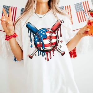 K&ouml;nnte beinhalten: Wei&szlig;es T-Shirt mit einem Baseball-Design, das die amerikanische Flagge enth&auml;lt, gekreuzt mit zwei Baseballschl&auml;gern. Der Baseball hat rote, wei&szlig;e und blaue Akzente mit einem tropfenden Farbeffekt. Die Schl&auml;ger sind blau und rot.