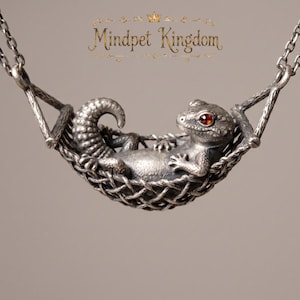 Op de afbeelding: Een zilverkleurige metalen hanger met een hagedis die in een geweven hangmat rust. De hagedis heeft een getextureerd lichaam en één rood edelstenen oog. Boven de hanger staat de tekst Mindpet Kingdom in een decoratief gouden lettertype.