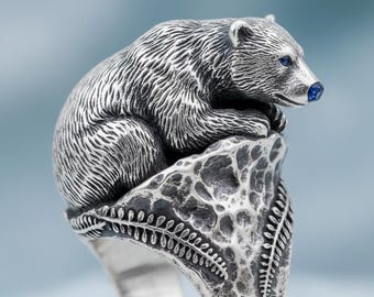 May include: A silver ring featuring a detailed bear design. The bear is perched atop a textured rock, with blue accents on its eyes and nose. The ring has a detailed, nature-inspired design.