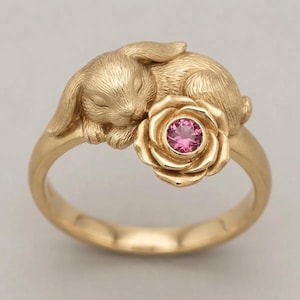 May include: A gold-coloured ring featuring a sleeping bunny resting on a rose. The rose has a pink gemstone centre. The ring's band is smooth and rounded, and the bunny and rose are intricately detailed.