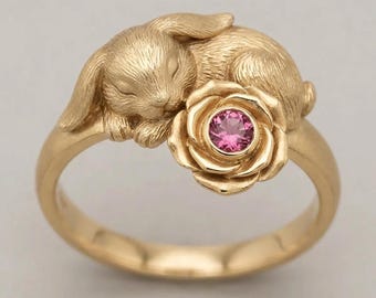 Könnte beinhalten: Ein goldener Ring mit einem skulptierten, schlafenden Kaninchen neben einer blühenden Rose. In der Mitte der Rose befindet sich ein kleiner, runder, facettierter rosa Edelstein. Das Metall hat eine glatte, polierte Oberfläche am Ringband.