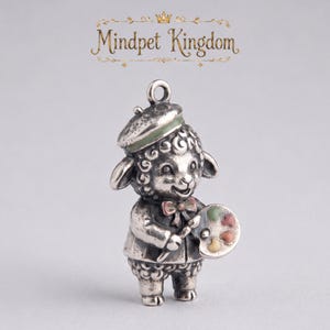 Könnte beinhalten: Ein silberfarbener Metallanhänger in Form eines Lamms mit Baskenmütze und Fliege, das einen Pinsel und eine Malerpalette mit kleinen farbigen Akzenten hält. Über der Lammfigur ist der Text Mindpet Kingdom zu sehen.