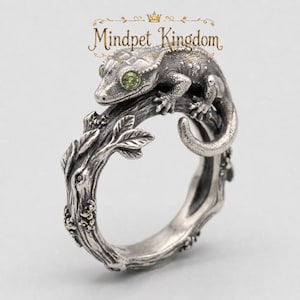Könnte beinhalten: Ein silberner Ring in Form eines strukturierten Baumastes mit Blättern und einem darauf ruhenden Gecko. Der Gecko hat zwei kleine, runde, hellgrüne Steine als Augen. Der Text Mindpet Kingdom steht in goldener Schrift über dem Ring.