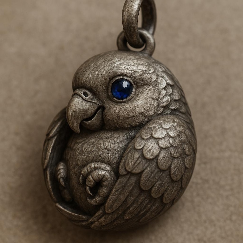 May include: A detailed silver-toned pendant in the shape of a small bird, possibly a parrot or parakeet. The bird has a blue gemstone eye and is curled up. The pendant has a loop at the top for a chain.
