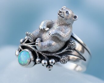 May include: A silver ring featuring a detailed bear figure reclining on a branch with small berries. The ring includes a round, opalescent stone. The ring has a wide band with a polished finish.