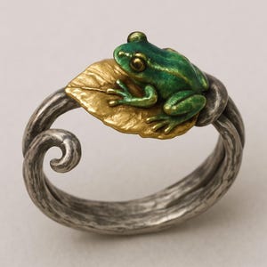 Könnte beinhalten: Ein silberfarbener Ring mit einer detaillierten grünen Froschfigur, die auf einem goldfarbenen Blatt sitzt. Das Ringband hat ein strukturiertes, gedrehtes Design. Der Frosch hat goldfarbene Augen und grüne und goldfarbene Akzente.