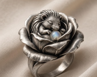 May include: A silver ring featuring a sculpted hedgehog resting inside a blooming rose. The hedgehog is curled up with its eyes closed, holding a small, round, iridescent moonstone bead between its front paws. The metal has a dark, oxidized finish.