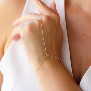 May include: A delicate gold hand chain featuring star-shaped charms. The chain connects to a ring and drapes across the back of the hand, with star accents along the chain. The jewelry is worn on a person's hand.