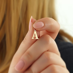 Könnte beinhalten: Ein goldfarbener Anhänger in Form des Buchstabens "A". Der Anhänger ist klein und hat oben eine Schlaufe zum Befestigen an einer Kette. Der Anhänger wird von den Fingern einer Person gehalten.