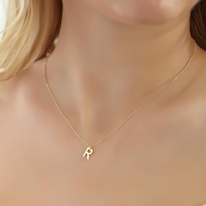 Könnte beinhalten: Eine zarte goldene Halskette mit einem Anhänger in Form des Buchstabens "R". Der Anhänger hängt an einer feinen Kette und erzeugt einen schlichten und eleganten Look. Die Halskette wird um den Hals getragen.
